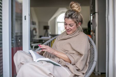 Young beautiful woman sits and reads a book on a terrace covered with a blanket and in the background there is a fireplaceの写真素材