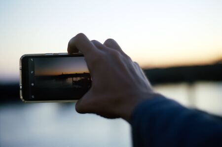 A hand extends out toward a sunrise in focus with a cell phone in the foreground, and is ready to take a picture of the horizon which is blurry in the background.の写真素材