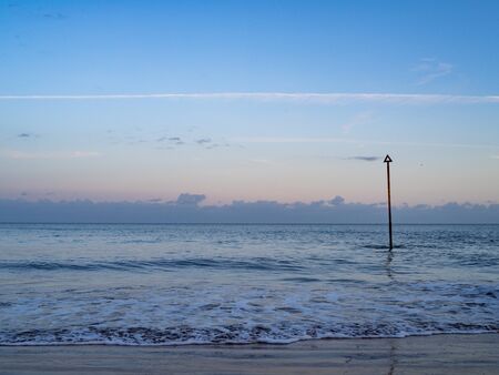A pole sticks up through the small waves on a beach with a soft blue sky.の写真素材
