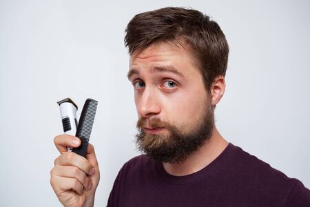 Young bearded man holding up an electrical trimmer and comb, keen to sort out his messy beard