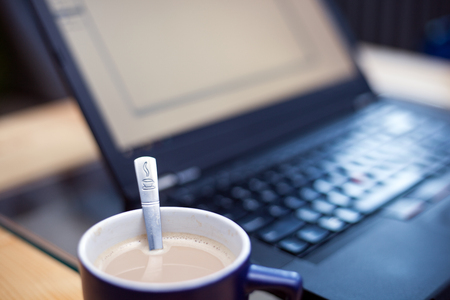 Fresh cup of coffee with a teaspoon stamped with a coffee symbol in focus with a laptop in the backgroundの写真素材