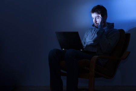 Tired and stressed young man working on a laptop in a dark room at nightの写真素材
