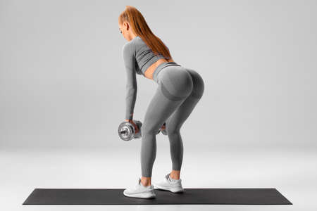Athletic girl doing deadlift exercise for glutes on gray background. Fitness woman working out with dumbbellsの写真素材