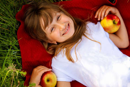 Nice happy little blonde girl with red apples, close-up. On the background of green grass.の写真素材