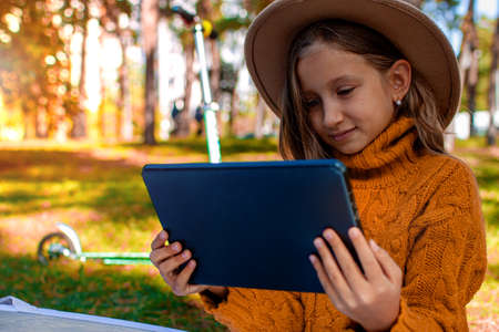 Little Girl Sitting In Park On Sunny Day And Play With Tablet Pc In Autumn Park.の写真素材
