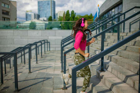 Waist up portrait of happy young asian woman training. She is listening to music from earphones and looking at smartphone. Athlete is standing outdoors and smiling.の写真素材