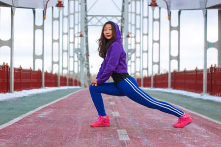 Athletic Asian girl in a hat and tracksuit runs on a bridge in the city. Healthy lifestyle and sports. Beautiful girl doing exercises in the winter.の写真素材