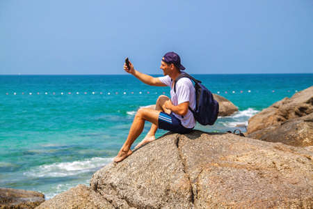 Side view of a happy man with backpack looking at sea while sitting on hill against sky during sunny day.の写真素材