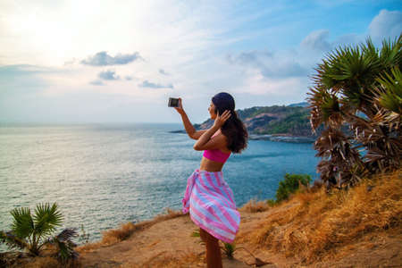 Asian Girl Tourist with backpack and a bottle of water standing on top of a hill with raised hands at sunset.の写真素材