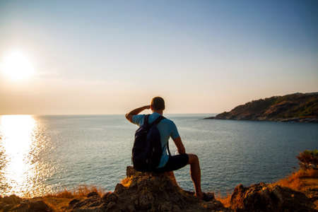 Side view of a happy man with backpack looking at sea while sitting on hill against sky during sunny day.の写真素材
