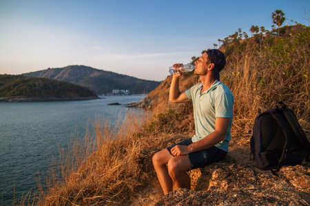 Side view of a happy man with backpack looking at sea while sitting on hill against sky during sunny day.の写真素材