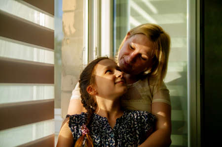 Mother and daughter sitting on sill near window in room. They show emotions and have fun.の写真素材