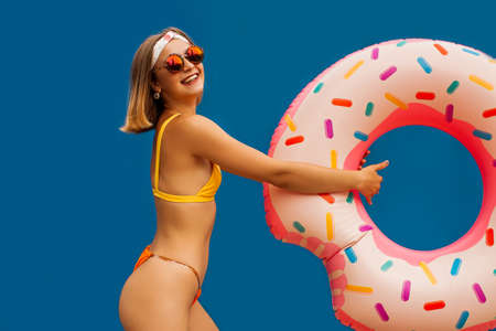 Beach Style. Blonde Woman Standing Isolated On Blue Wall Posing To Camera With Swim Inflatable Ring.