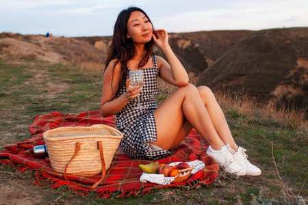 Mature Asian Woman Holding Glass Of Wine Watching Sunset Over Hills On Summer Picnic.の写真素材