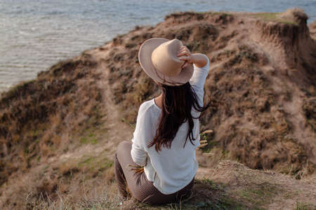 Young Slender Woman In A Hat Sits On A Cliff On A Background Of Blue Sea.