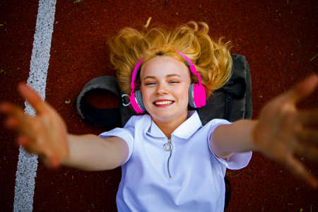 Top View Of Young Blonde Teen Girl With Headphone Lying On Backpack Outdoor.