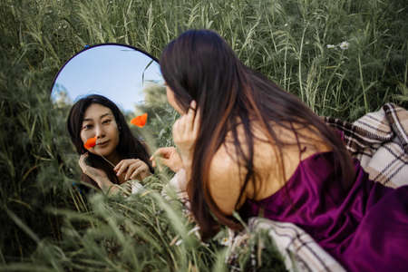 Mirror Reflection Of Young Pretty Asian Woman Holding Red Poppy Flower In Green Fieldの写真素材