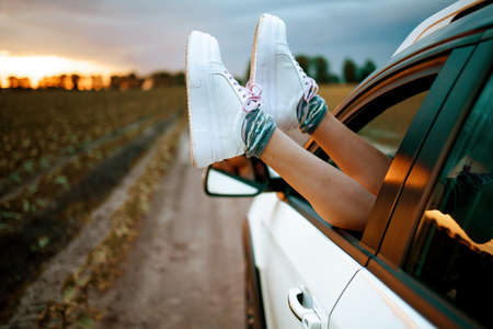 Woman Feet In Sneakers On Car Window At Sunset. Roadtrip And Freedom Travel Concep