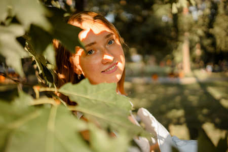 Portrait Of A Young Red Hair Woman With Face Obscured By Green Leaves In Forest.の写真素材
