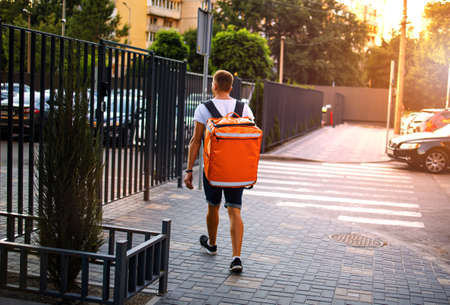 Young deliveryboy walking with orange thermal bag on city street. Man of delivery service in hurry to deliver an order. Delivery service goes to give the order quickly to the clientの写真素材