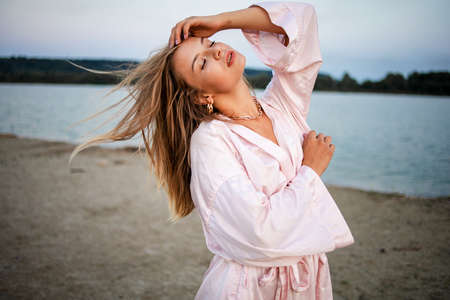 Romantic blonde woman in pink robe on the beach by the sea at sunset.の写真素材