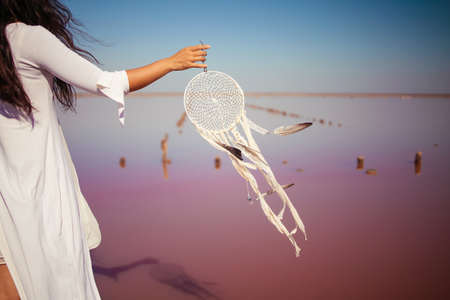 Photo of beautiful boho styled model wearing white on the beach of pink lake in sunset. Dream catchers blown around by a light sea breezeの写真素材