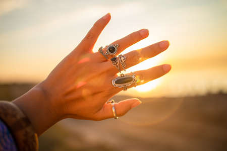 Hippie woman with dark long hair, silver rings with stone and white blouse stands back at sunset. Indie boho vibes and bohemian styleの写真素材