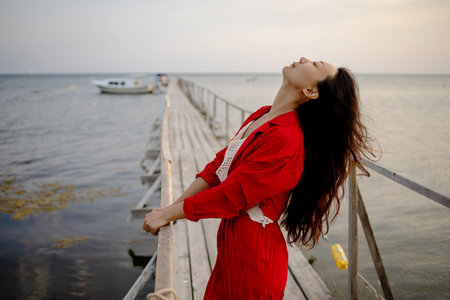 Asian Girl in red standing with open arms on wooden bridge against sky, seagulls fly over the beach. Freedom conceptの写真素材