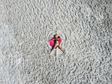 Summer holiday - tanning girl wearing sun hat at the beach on a white sand shot from above. Top view from drone. Aerial view of slim woman sunbathing lying on beach.の写真素材