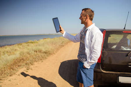 young caucasian man leaning against a car working on a tablet, communicates on a video call.