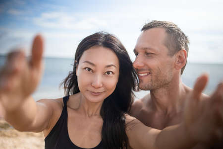 Image of young happy man kissing and hugging beautiful asian woman while taking selfie photo on sunny beachの写真素材
