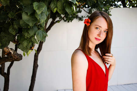 Portrait of a beautiful blonde woman in red dress outdoor near big tree and white wallの写真素材
