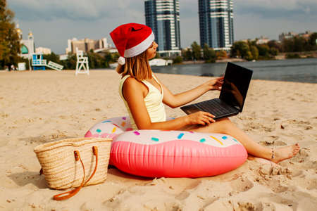 Pretty woman in Santa hat at tropical ocean beach using laptop computer, online chooses gifts for the new yearの写真素材