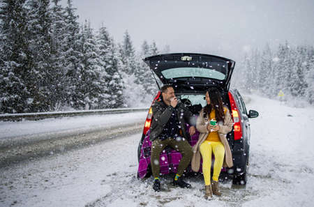 outdoor fashion portrait of young mixed race couple sitting in car trunk under falling snow in cold winter forest.の写真素材