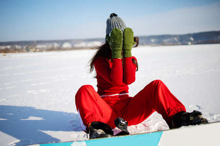 Asian woman fitness model in a red swimsuit, snowboard boots smiling, stands on a snowboard and attractive poses on a ski slope in winter, rear view, in the background forestの写真素材