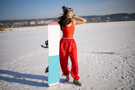 Asian woman fitness model in a red swimsuit, snowboard boots smiling, stands on a snowboard and attractive poses on a ski slope in winter, rear view, in the background forestの写真素材