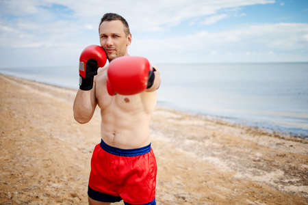 young male bodybuilder shows athletic figure strong torso at sunset on beach. sexy boxers training exercise.の写真素材