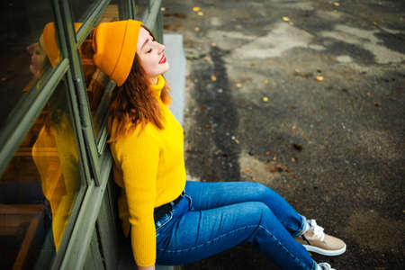 Portrait of fashionable blonde girl with red lipstick wearing yellow sweater and hat outdoors in the cityの写真素材