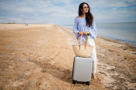 Young woman with her suitcase luggage on beautiful sandy beach view on sunny day. Asian female on ocean shore having fun watching waves, sunbathing. Background, close upの写真素材