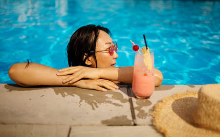 slim young asian woman in swimming pool with glass of juice, relax and feel comfortable life in summer time holiday and Vacationの写真素材