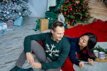Happy charming asian woman dressed in jersey talking to her handsome boyfriend spending time in a cozy home, romantic couple in love happy about celebrating winter holidays together in good mood.の写真素材
