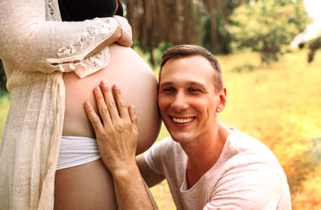 Young Happy Family, handsome husband kissing newborn baby through wife belly in nature.