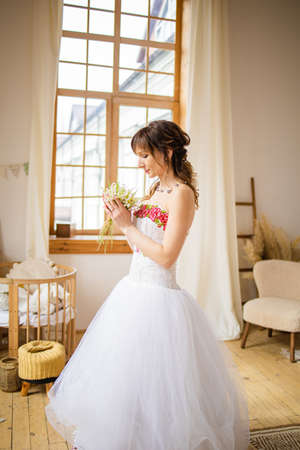Portrait of the bride in a lush wedding dress and hairstyle, she near big window waiting for the groom.の写真素材