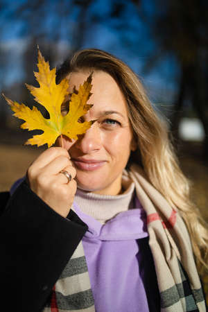 Beautiful happy woman with a smile holds an autumn yellow leaf near the faceの写真素材
