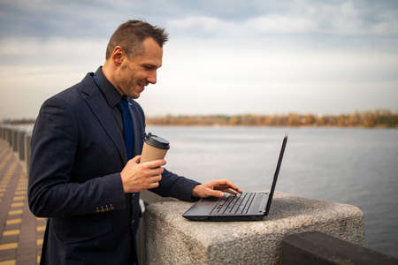 Young handsome businessman standing next to his laptop on the bridge near lake.