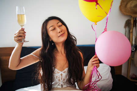 Smiling pretty young asian woman holds bundles of balloons and champagne on bed in morning.の写真素材