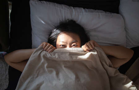 Beautiful asian woman is Waking up in the Morning, Stretches in the Bed, Sun Shines on Her From Window. Happy Young Girl Greets New Day with Warm Sunlight Flare.の写真素材