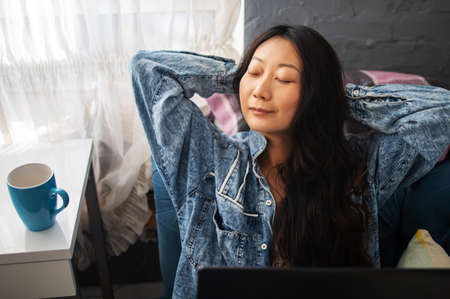 positive young asian woman resting her hands behind her head after a long study on a laptop at a large home table in a modern apartmentの写真素材