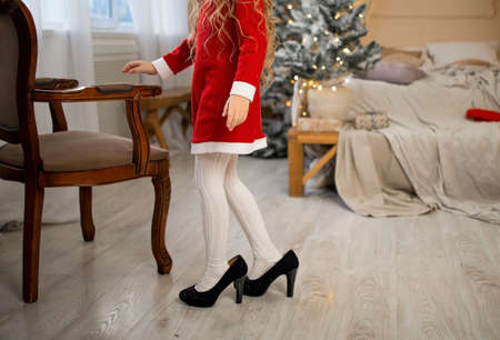 Cute little girl in red Santa dress. Happy child girl try on outfits and mom's shoes in Christmas eve.の写真素材