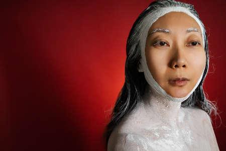 clay mask on face, enjoys softness, has beauty treatments at home, poses half naked, isolated over red background. Skin care concept.の写真素材